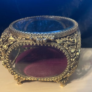 May include: Ornate gold-toned jewelry box with a clear glass top and sides. The box features intricate floral and scrollwork detailing. The interior is lined with a deep purple velvet. The box is raised on small, decorative feet.