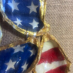 Puede incluir: Primer plano de varias conchas de ostra decoradas con dise&ntilde;os patri&oacute;ticos. Algunas conchas presentan un fondo azul con estrellas blancas, mientras que otras muestran rayas rojas y blancas, todas con bordes pintados de oro. Las conchas est&aacute;n dispuestas sobre una superficie texturizada y beige.