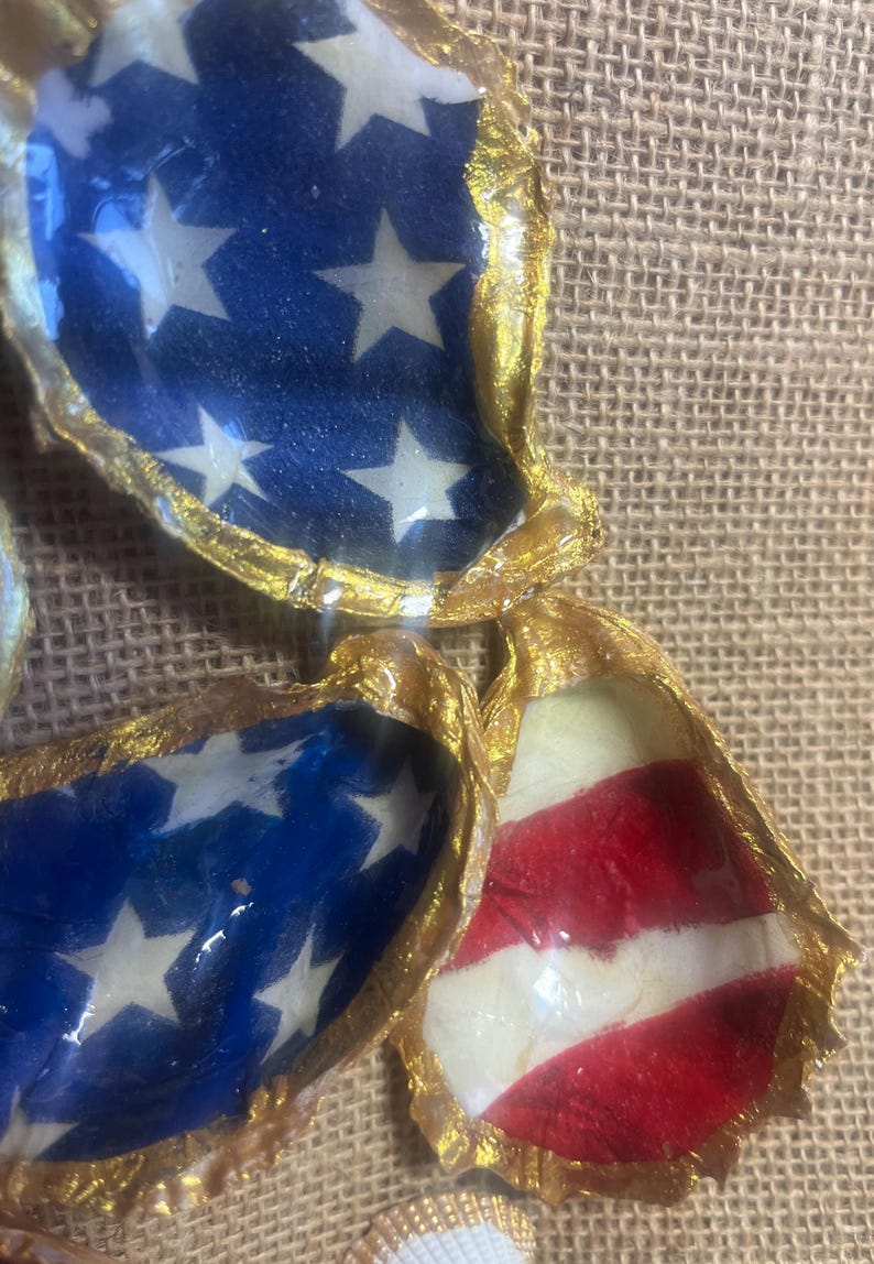 Puede incluir: Primer plano de varias conchas de ostra decoradas con dise&ntilde;os patri&oacute;ticos. Algunas conchas presentan un fondo azul con estrellas blancas, mientras que otras muestran rayas rojas y blancas, todas con bordes pintados de oro. Las conchas est&aacute;n dispuestas sobre una superficie texturizada y beige.