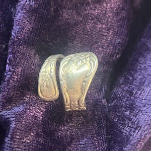 May include: A silver-toned ring made from repurposed silverware, featuring floral and scrollwork details. The ring is set against a deep purple, textured background, showcasing its vintage aesthetic. The ring's design is a unique, handcrafted piece.