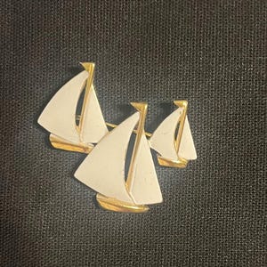 May include: A gold-toned brooch featuring three sailboats with white sails and gold accents. The sailboats are arranged in a cluster, with the sails angled to catch the wind. The brooch is set against a dark background.