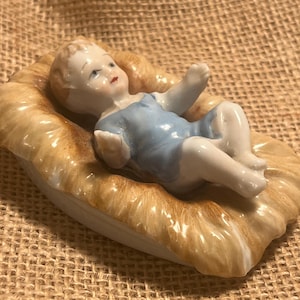 May include: A ceramic figurine of a baby in a light blue outfit, lying in a bed of straw. The figurine has detailed facial features and is resting on a white base with a straw-like texture.