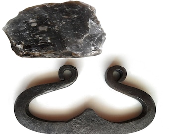 May include: A dark, textured flint rock and a dark metal fire starter. The fire starter is a curved, hook-shaped tool with a central notch and looped ends. The flint is a dark gray color with lighter inclusions.