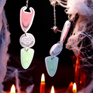 Puede incluir: Un par de pendientes colgantes con temática de Halloween. Los pendientes presentan una cara de diablo roja, una cara de calavera blanca y una cara triste verde. Los pendientes cuelgan de cadenas plateadas.