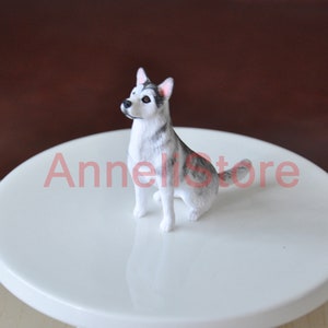 May include: A small, detailed figurine of a sitting husky dog with grey and white fur, pointed ears, and dark eyes. The dog is positioned in a seated pose with its head turned slightly to the side. The text Annelistore is visible in red.