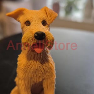 May include: A tan-colored dog figurine with textured fur, dark brown eyes, a black nose, and a small pink tongue visible between its mouth. The text "AngelStore" is overlaid in red across the center of the figurine's face and chest area.