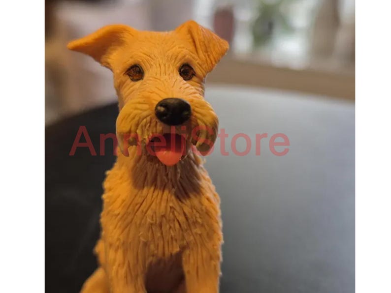 May include: A tan-colored dog figurine with textured fur, dark brown eyes, a black nose, and a small pink tongue visible between its mouth. The text "AngelStore" is overlaid in red across the center of the figurine's face and chest area.