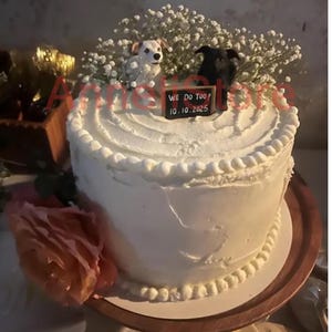 May include: A round white frosted cake topped with white baby's breath flowers, a small black and white dog figurine, and a black rectangular sign with the white text WE DO TOO! 10.10.2025. A large peach-colored artificial flower is attached to the side.