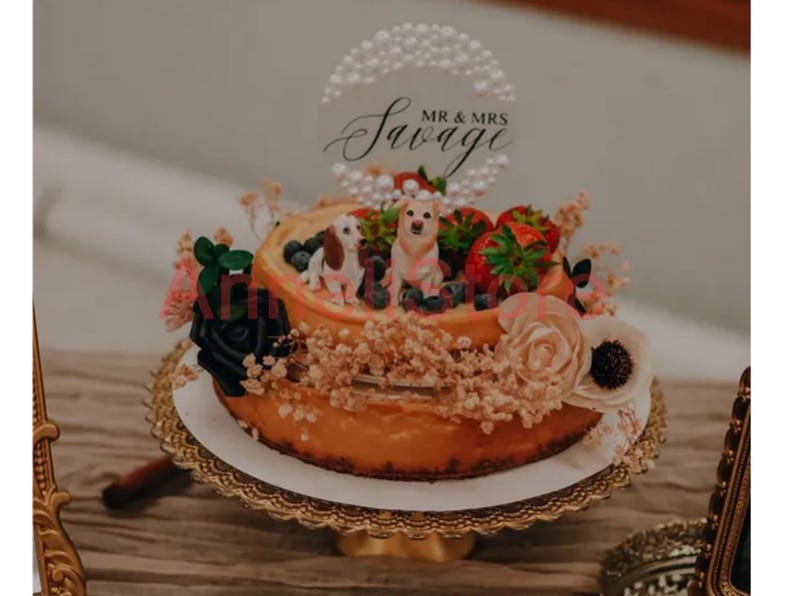 May include: A two-tiered cake topped with two dog figurines, fresh strawberries, and blueberries. A circular acrylic cake topper features a pearl border and the black script text MR & MRS Savage. The cake is decorated with black and cream flowers.