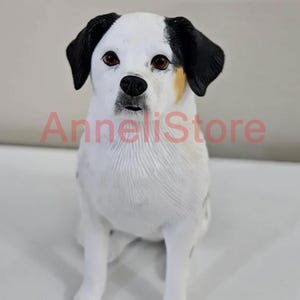 May include: A dog figurine with white fur, black ears, and brown eyes, featuring a small patch of tan fur near the left ear. The text AnneliStore is overlaid in red across the center of the figurine, which is depicted in a seated position.