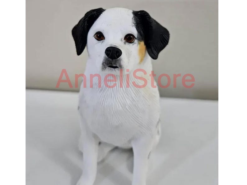 May include: A dog figurine with white fur, black ears, and brown eyes, featuring a small patch of tan fur near the left ear. The text AnneliStore is overlaid in red across the center of the figurine, which is depicted in a seated position.