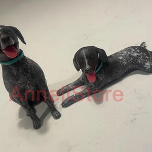 May include: Two dog figurines with black and white speckled coats, teal collars, and open mouths with red tongues. One dog is in a sitting position, while the other is lying down. The text "AnneliStore" is visible in red across the center.