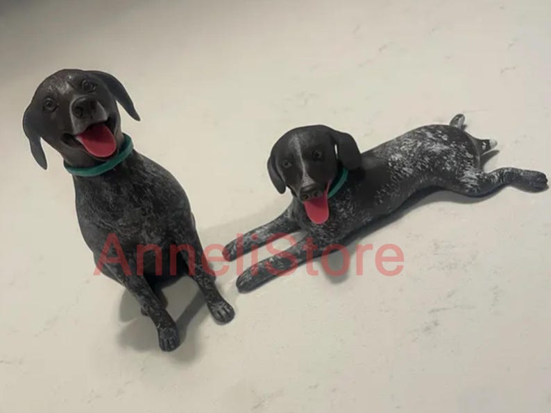 May include: Two dog figurines with black and white speckled coats, teal collars, and open mouths with red tongues. One dog is in a sitting position, while the other is lying down. The text "AnneliStore" is visible in red across the center.