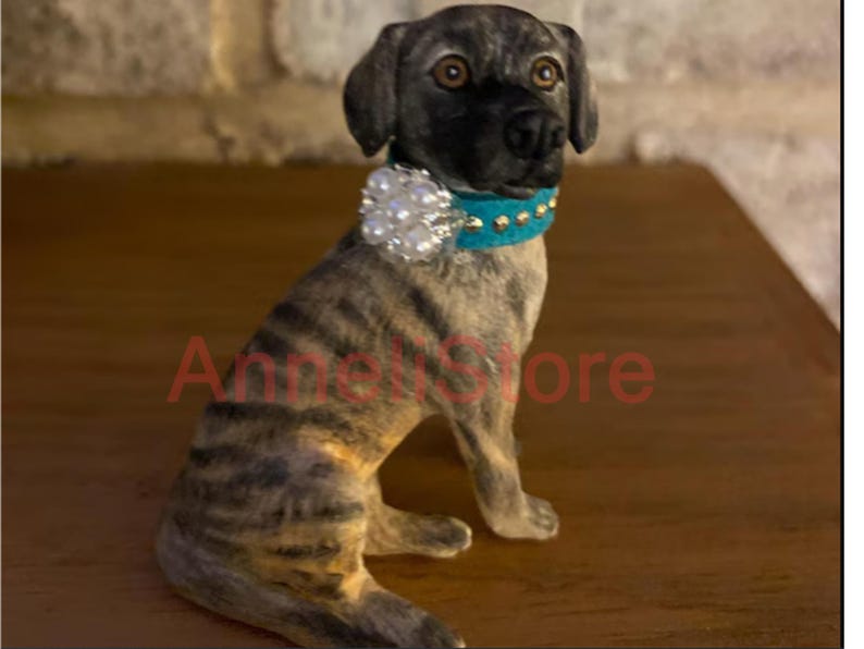 May include: A brindle dog figurine with a dark muzzle and brown eyes, wearing a bright turquoise collar adorned with gold-tone studs and a large, round cluster of white faux pearls. The text AnneliStore is visible in red across the dog's body.