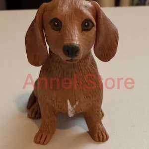 May include: A brown dachshund figurine with long floppy ears, dark eyes, and a black nose. The dog is in a sitting position with a small white mark on its chest. The text "AnneliStore" is superimposed in red across the center of the figurine.