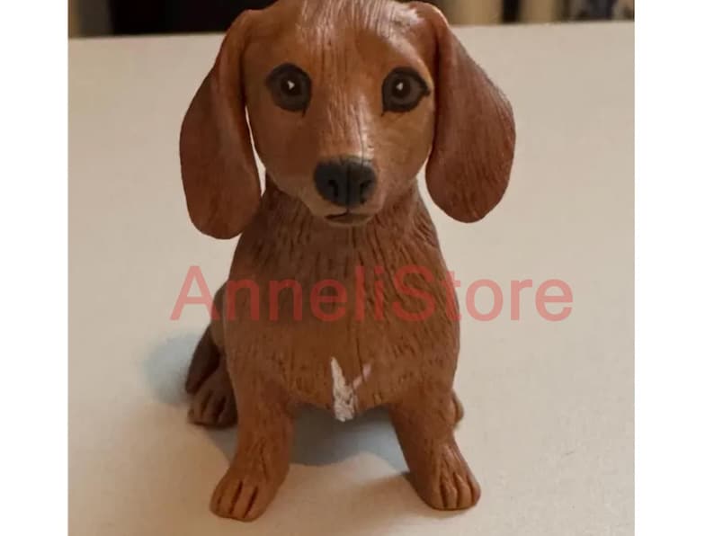 May include: A brown dachshund figurine with long floppy ears, dark eyes, and a black nose. The dog is in a sitting position with a small white mark on its chest. The text "AnneliStore" is superimposed in red across the center of the figurine.