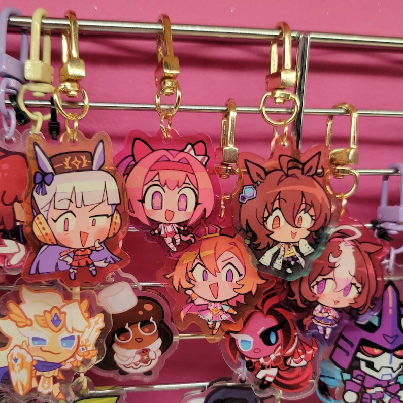K&ouml;nnte beinhalten: Eine Sammlung farbenfroher Schl&uuml;sselanh&auml;nger mit Anime-Charakteren, die an einem Metallgitter h&auml;ngen. Die Schl&uuml;sselanh&auml;nger zeigen verschiedene Charaktere in leuchtenden Farben, darunter Pink, Lila, Orange und Rot. Jeder Schl&uuml;sselanh&auml;nger hat einen goldfarbenen Verschluss.