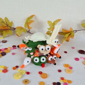Può includere: Una collezione di piccole figure di animali all'uncinetto impilate insieme, tra cui una rana verde, un orso beige e un coniglio bianco con lunghe orecchie. Le figure presentano occhi in plastica nera e sono circondate da bottoni sparsi.