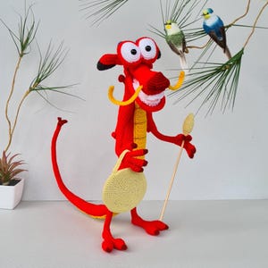 Op de afbeelding: Een handgemaakte gehaakte draak in helder rood, geel en wit. De draak heeft grote ogen, gele hoorns en een gele buidel. Hij houdt een houten stok met een gele bovenkant vast. Decoratieve vogels en groen zijn op de achtergrond.