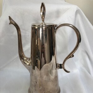 Puede incluir: Una cafetera de plata con un cuerpo alto y delgado y un asa curva. La cafetera tiene un acabado pulido y un remate decorativo en la tapa.