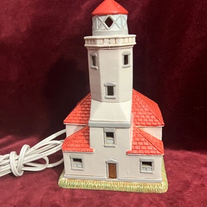 Puede incluir: Un faro de cerámica blanca con techo rojo y base blanca. El faro tiene una luz en la parte superior y ventanas en los lados. El faro está sentado sobre una base verde.