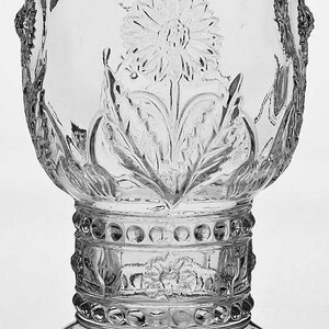 May include: Clear glass goblet with a floral design. The goblet features an embossed flower and leaf pattern, with decorative detailing around the base and stem. The glass has a rounded bowl shape and a sturdy base.