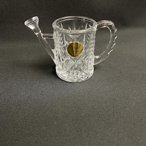 May include: Clear glass watering can with a diamond pattern and a handle. The watering can has a spout and a gold sticker on the front. The watering can is a decorative item.