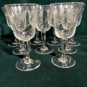 May include: Set of eight clear glass stemmed drinking glasses with a delicate etched design. The glasses are arranged in a circular pattern on a dark green surface.