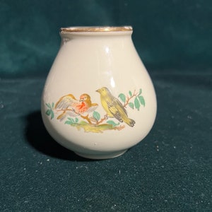 Puede incluir: Un pequeño jarrón de cerámica blanca con un borde dorado, que presenta un diseño pintado de dos pájaros posados en una rama con hojas verdes.