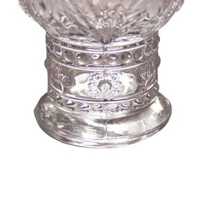 May include: A clear, ornate glass vase with a wide base and a detailed, textured design. The vase features a series of decorative bands and floral patterns, creating a vintage aesthetic. The glass reflects light, highlighting the intricate details.