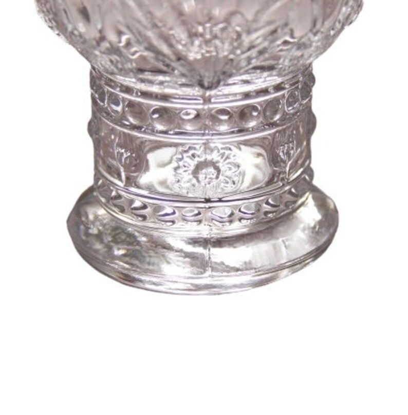 May include: A clear, ornate glass vase with a wide base and a detailed, textured design. The vase features a series of decorative bands and floral patterns, creating a vintage aesthetic. The glass reflects light, highlighting the intricate details.