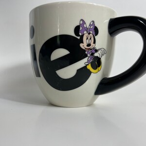 Puede incluir: Taza de cer&aacute;mica blanca con asa negra. La taza tiene un gr&aacute;fico en blanco y negro de la palabra "ie" con Minnie Mouse sentada en la "e". Minnie Mouse lleva un vestido morado con lunares blancos y un lazo rojo.