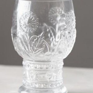 May include: Clear glass goblet with a detailed embossed floral design. The glass features a wide bowl, a decorative stem, and a round base. The floral pattern includes dandelions and leaves, adding a touch of nature to the glassware.