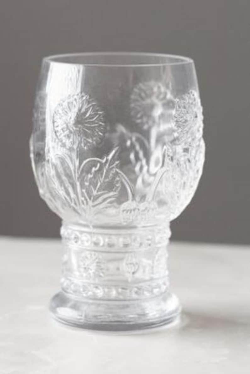 May include: Clear glass goblet with a detailed embossed floral design. The glass features a wide bowl, a decorative stem, and a round base. The floral pattern includes dandelions and leaves, adding a touch of nature to the glassware.