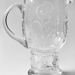 May include: Clear glass pitcher with a large handle and a floral design. The pitcher features an embossed dandelion and leaf pattern. The base has a decorative beaded design. The pitcher is ideal for serving beverages.