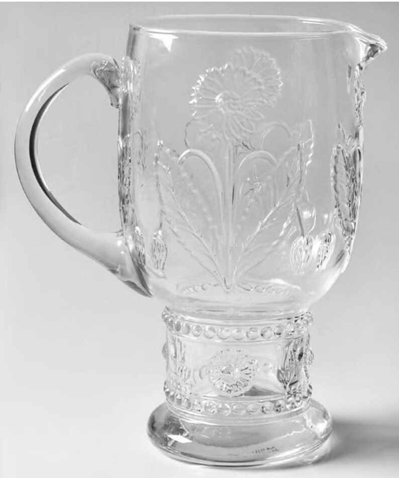 May include: Clear glass pitcher with a large handle and a floral design. The pitcher features an embossed dandelion and leaf pattern. The base has a decorative beaded design. The pitcher is ideal for serving beverages.