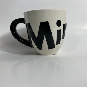 Puede incluir: Taza de cer&aacute;mica blanca con asa negra y la palabra "Mir" en letras negras grandes.
