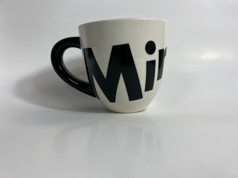Puede incluir: Taza de cer&aacute;mica blanca con asa negra y la palabra "Mir" en letras negras grandes.
