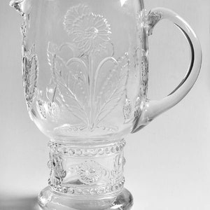 May include: Clear glass pitcher with a large handle and a spout. The pitcher features embossed floral and leaf designs, with a prominent flower in the center. The base has a decorative band with a beaded design.