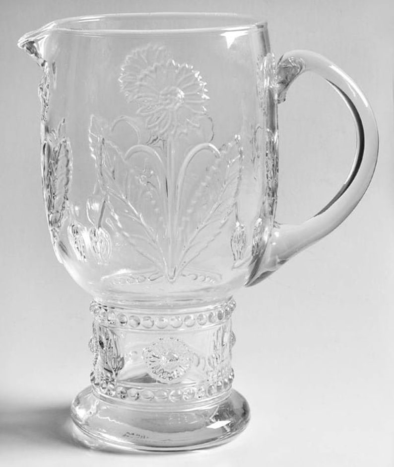 May include: Clear glass pitcher with a large handle and a spout. The pitcher features embossed floral and leaf designs, with a prominent flower in the center. The base has a decorative band with a beaded design.