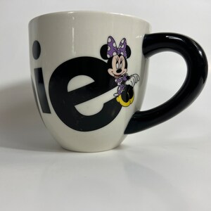 Puede incluir: Taza de cer&aacute;mica blanca con asa negra. La taza tiene un gr&aacute;fico en blanco y negro de la palabra "ie" con Minnie Mouse sentada en la "e". Minnie Mouse lleva un vestido morado con lunares blancos y un lazo rojo.