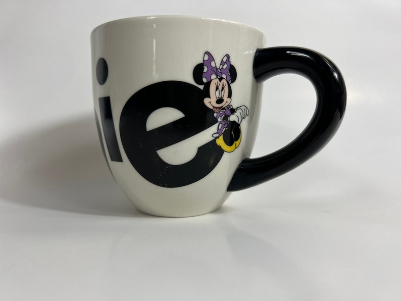 Puede incluir: Taza de cer&aacute;mica blanca con asa negra. La taza tiene un gr&aacute;fico en blanco y negro de la palabra "ie" con Minnie Mouse sentada en la "e". Minnie Mouse lleva un vestido morado con lunares blancos y un lazo rojo.