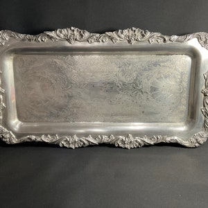 May include: A silver serving tray with ornate handles and a decorative embossed design. The tray is rectangular and has a slightly curved edge.