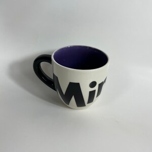 Puede incluir: Taza de cer&aacute;mica blanca con asa negra y la palabra "Mir" en letras negras en la parte delantera. El interior de la taza es morado.