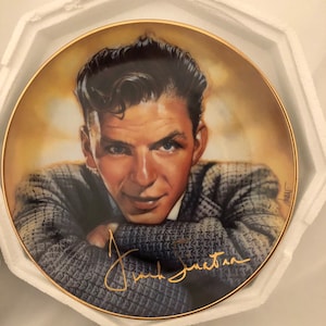 Peut inclure: Une assiette en porcelaine représentant un portrait de Frank Sinatra portant un pull bleu et gris. L'assiette a un bord doré et une signature dorée de Frank Sinatra.
