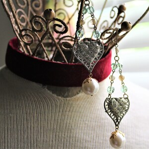 May include: Two silver heart-shaped pendants with rhinestones and pearl accents hang from a wire stand. The pendants are attached to chains with green and white beads.