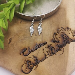 Könnte beinhalten: Silberne Muschel-Ohrringe mit einem zarten Design. Die Ohrringe hängen an einer Holzoberfläche mit dem eingravierten Text "Christmas Schmuck Dave".