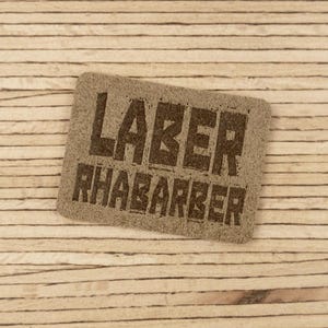 Könnte beinhalten: Ein rechteckiges, hellbraunes Etikett mit den Worten "LABER RHABARBER" in dunkelbraunen Blockbuchstaben. Das Etikett befindet sich auf einem hellbraunen Hintergrund mit Holzmaserung.