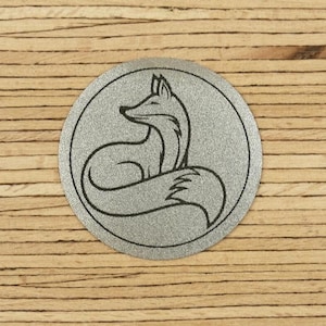Könnte beinhalten: Ein runder, silberfarbener Aufnäher mit einem stilisierten Fuchs-Design. Der Fuchs ist in einer sitzenden Position dargestellt, mit dem Schwanz um seinen Körper gelegt. Der Fuchs ist schwarz umrandet, auf Holzgrund.