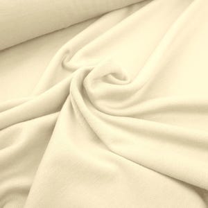 Könnte beinhalten: Nahaufnahme eines cremefarbenen Stoffes mit weicher, strukturierter Oberfläche. Der Stoff ist drapiert und gewirbelt, wodurch Falten und Schatten entstehen. Das Material scheint ein Strick- oder Jersey-Stoff zu sein, geeignet für Kleidung oder Textilprojekte.